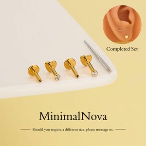 Puede incluir: Un conjunto de pendientes o piercings labiales dorados con varios diseños, incluyendo formas de corazón y tapas redondas, algunos con piedras preciosas transparentes. También se incluye una herramienta de perforación plateada. La imagen también muestra un primer plano de una oreja con un pendiente.