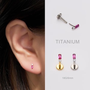 May include: Titanium ear piercing studs with a rectangular pink gemstone and a small white gemstone. The image shows the stud in the ear, and two other studs with gold and silver bases. The text "TITANIUM" is also visible.
