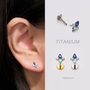 May include: An ear with a small, blue and white gemstone earring is shown. The image also shows two other earrings with blue and white gemstones, and the word "TITANIUM". The earrings are 6mm in size.