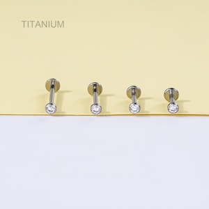 May include: Four titanium labret studs with clear, round cubic zirconia stones. The studs are arranged in a row against a yellow and white background. The word "TITANIUM" is printed in the upper left corner.