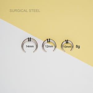 May include: Three silver surgical steel septum rings with black o-rings. The rings are 14mm, 12mm, and 10mm in diameter. The rings are labeled with their size in millimeters. The text "SURGICAL STEEL" is at the top of the image. The text "8g" is at the right side of the image.