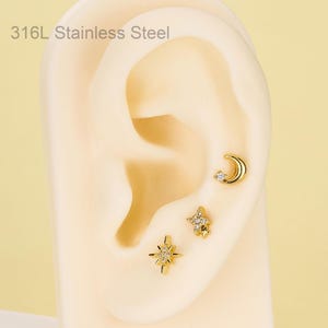 May include: Three gold-tone 316L stainless steel stud earrings featuring celestial motifs. The set includes a crescent moon with a small clear stone, a four-pointed star with multiple clear stones, and a larger eight-pointed star with clear stones.