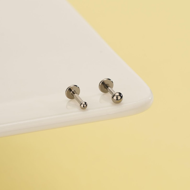 Pode incluir: Dois brincos de l&aacute;bio ou orelha prateados. Cada brinco tem uma pequena bola redonda na extremidade de um pino. Os brincos s&atilde;o exibidos em uma superf&iacute;cie branca com um fundo amarelo.