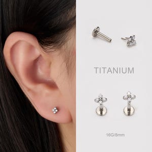 May include: A close-up of an ear with a small, silver stud earring. The earring is a flower shape with clear, sparkling stones. Additional earrings and the word "TITANIUM" are also visible.