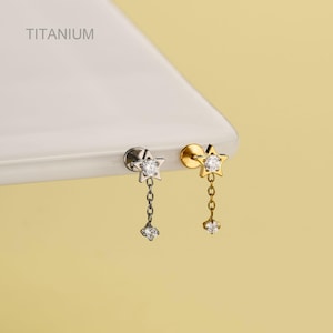 May include: Two star-shaped earrings with dangling charms. One earring is silver-toned, the other gold-toned. Each has a clear gem in the center of the star and at the end of the chain. The word "TITANIUM" is in the upper left corner.