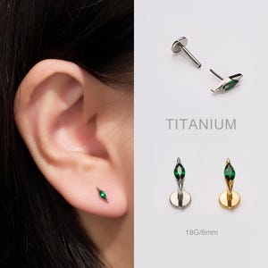 May include: An ear with a small, silver stud earring with a green gemstone. The image also shows two additional stud earrings with green gemstones, one silver and one gold. The text "TITANIUM" is visible. The earrings are 6mm in length.