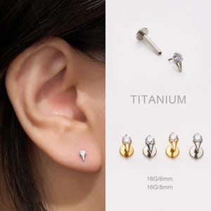 Puede incluir: Primer plano de una oreja con un pequeño pendiente de titanio plateado con un diseño en forma de cono y una gema transparente. Se muestran pendientes adicionales con bases doradas y plateadas. También se ve el texto "TITANIUM" y "16G/6mm 16G/8mm".