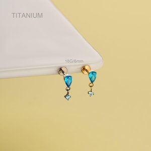 May include: A pair of earrings featuring a light blue, teardrop-shaped gemstone suspended from a small stud. The earrings are displayed against a white surface, with the text "TITANIUM" and size "18G/6mm" visible.