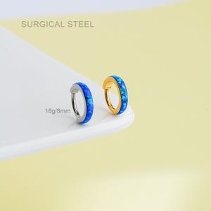 Może przedstawiać: Dwa kolczyki koła z niebieskimi inkrustacjami z opalu. Jeden kolczyk jest w kolorze srebrnym, a drugi w kolorze złotym. Tekst "SURGICAL STEEL" znajduje się na górze, a "16g/8mm" na dole. Kolczyki leżą na białej powierzchni na żółtym tle.