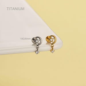 May include: Two earrings, one silver and one gold, each with a crescent moon and star design and a dangling clear gem. The earrings are labelled "TITANIUM" and "18G/6mm".