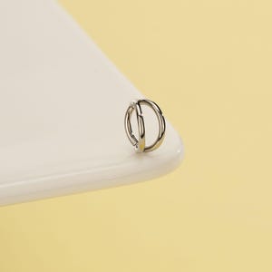 Pode incluir: Um brinco prateado de argola dupla. O brinco &eacute; um design pequeno e circular com duas argolas conectadas. A joia est&aacute; colocada sobre uma superf&iacute;cie branca com um fundo amarelo.