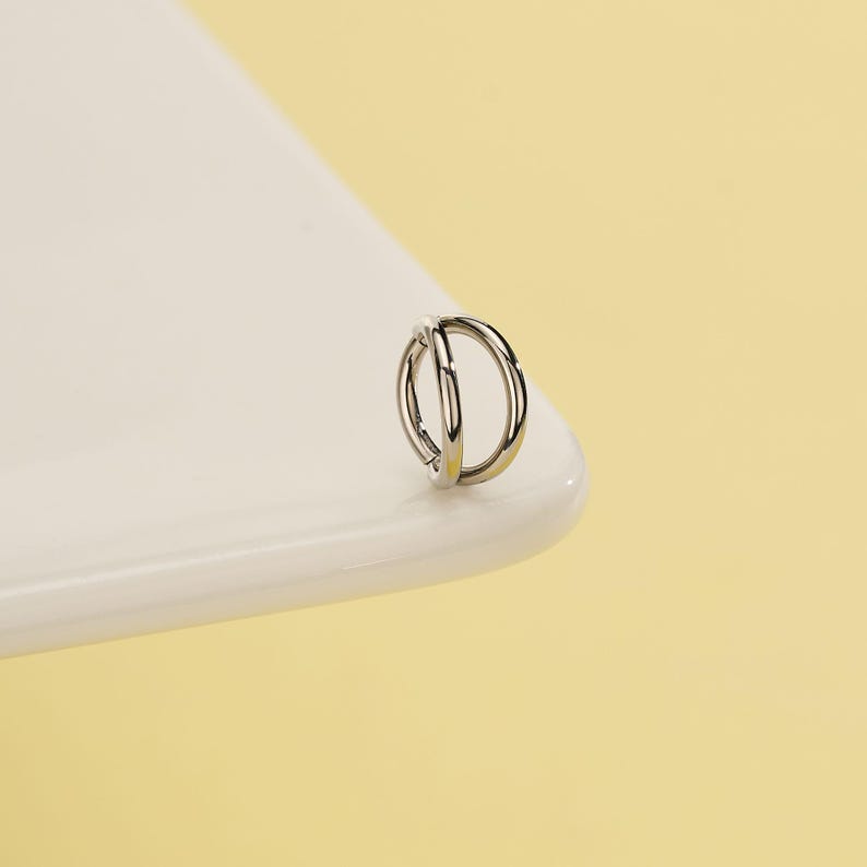 Pode incluir: Um brinco prateado de argola dupla. O brinco &eacute; um design pequeno e circular com duas argolas conectadas. A joia est&aacute; colocada sobre uma superf&iacute;cie branca com um fundo amarelo.