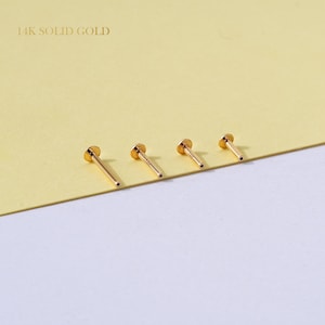 May include: Four 14K solid gold flat back labret studs arranged in a row. Each piece features a thin, straight post with a small, circular flat disc at one end. The metal has a polished, reflective yellow gold finish, and the text 14K SOLID GOLD is visible.