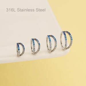 May include: Four stainless steel hoop earrings with a double-band design. Each earring features a gradient of blue and clear stones. The text "316L Stainless Steel" is visible in the image.