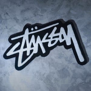 Könnte beinhalten: Ein weißer Stussy-Logo-Teppich mit schwarzem Umriss. Der Teppich besteht aus einem weichen, flauschigen Material.