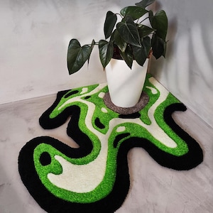 Könnte beinhalten: Ein abstrakt geformter Teppich mit einem auffälligen Design. Der Teppich hat einen schwarzen Rand, eine leuchtend grüne Mitte und weiße Akzente. Ein weißer Blumentopf mit dunkelgrünen Blättern steht auf dem Teppich und bringt Natürlichkeit in den Raum.