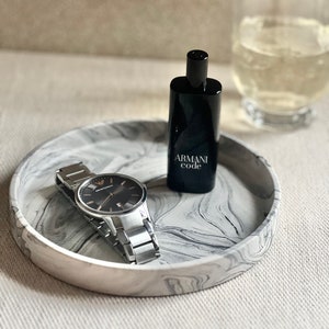 Op de afbeelding: Een zwart en zilveren horloge en een zwart flesje Armani Code cologne op een wit en grijs marmeren dienblad.