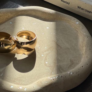 May include: A gold-colored pair of hoop earrings rests on a beige, organically shaped tray. The tray has a textured surface with small, white flecks. A book titled "Wasted" by Katie Treggiden is partially visible in the background.