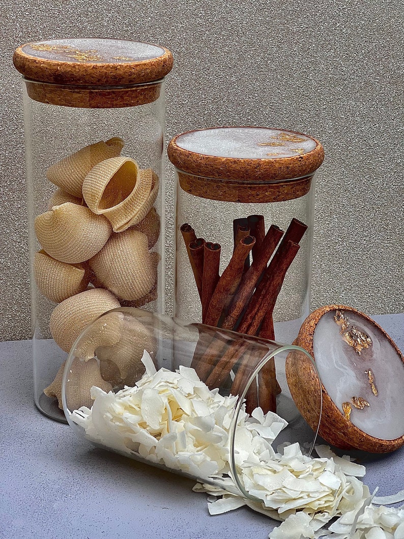 May include: Three glass jars with cork lids and gold accents. The jars are filled with various items, including pasta shells, cinnamon sticks, and shredded coconut.