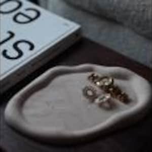 May include: A light brown, organic-shaped tray holds jewelry, including a gold watch and earrings. The tray sits on a dark wooden surface next to a book with the word "sea" visible on the cover.