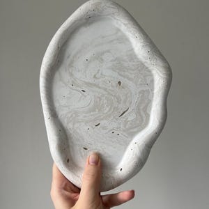 Peut inclure: Un plateau en béton fait à la main, de forme organique, dans des tons de blanc et de gris. Le plateau présente un motif marbré et une surface texturée. Ce plateau décoratif est idéal pour exposer des bijoux, de petits objets ou comme élément de décoration unique.