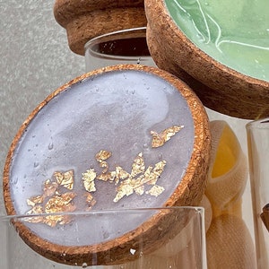 May include: Three glass jars with cork lids. The lids are decorated with resin and gold leaf. The resin is clear and has a marbled effect. The gold leaf is scattered randomly on the resin. The jars are empty.