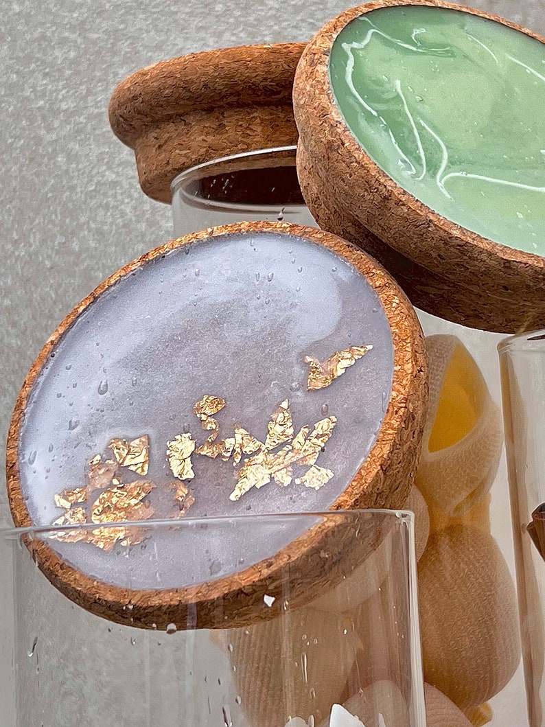 May include: Three glass jars with cork lids. The lids are decorated with resin and gold leaf. The resin is clear and has a marbled effect. The gold leaf is scattered randomly on the resin. The jars are empty.