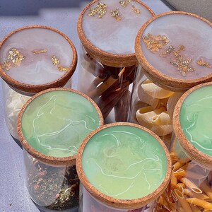 May include: Six glass jars with cork lids, each filled with different ingredients. The lids are decorated with a white and green marbled resin and gold leaf accents. The jars contain cinnamon sticks, pasta, dried herbs, and red pepper flakes.