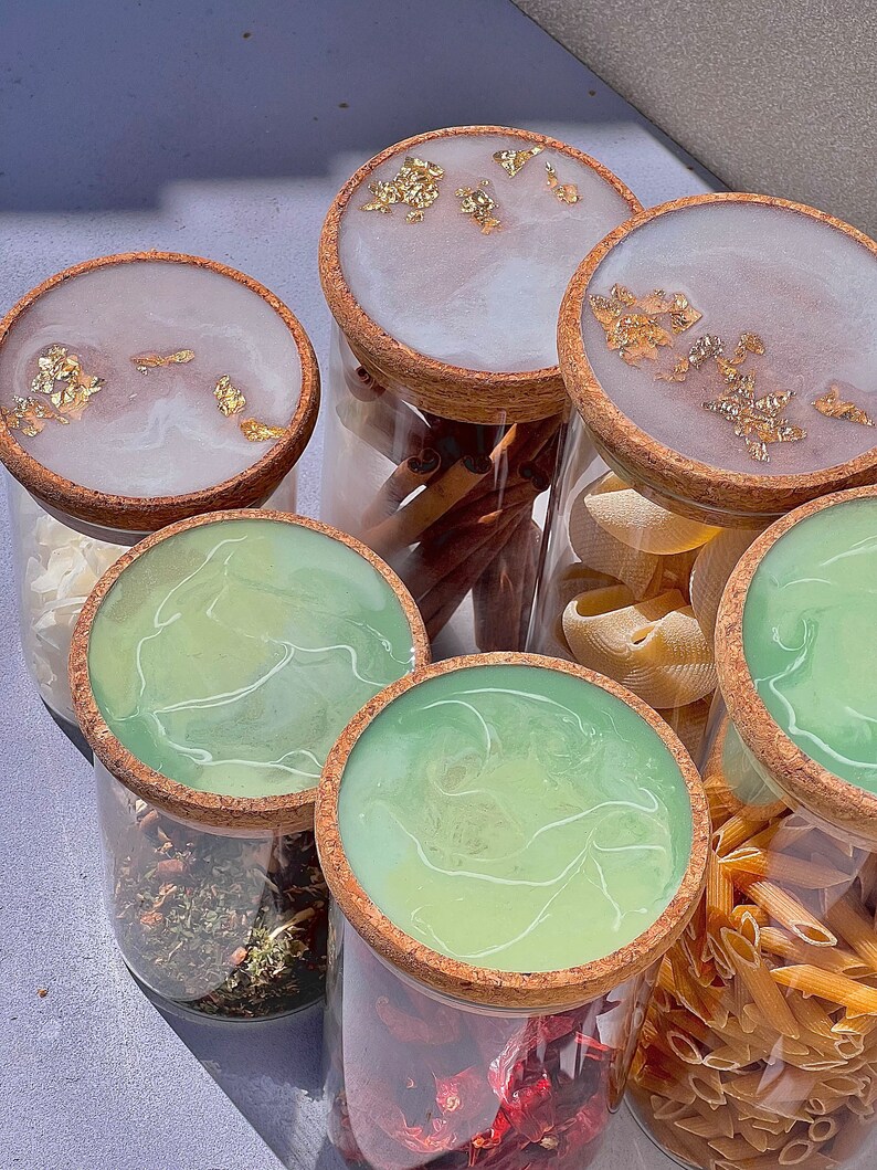 May include: Six glass jars with cork lids, each filled with different ingredients. The lids are decorated with a white and green marbled resin and gold leaf accents. The jars contain cinnamon sticks, pasta, dried herbs, and red pepper flakes.