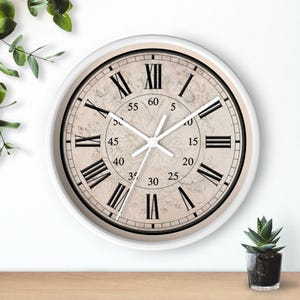 May include: A round wall clock with a white frame and a beige face featuring black Roman numerals for the hours and a central ring with black Arabic numerals from 5 to 60 for the minutes. The clock has white hands and a faint botanical illustration.