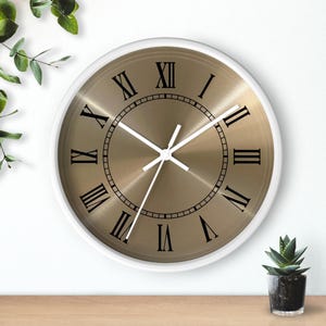 Könnte beinhalten: Eine runde Wanduhr mit weißem Rahmen und metallisch goldfarbenem Zifferblatt. Die Uhr verfügt über schwarze römische Ziffern als Stundenmarkierungen, eine dünne schwarze kreisförmige Minutenskala und drei weiße Zeiger auf der gebürsteten Goldfläche.