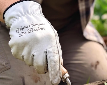 Puede incluir: Un par de guantes de jardinería de cuero blanco con ribetes negros y el texto "Marie Semeuse De Banheur" impreso en letra cursiva negra en el dorso de cada mano. Los guantes se usan mientras se utiliza una pala de mano para plantar un brote.