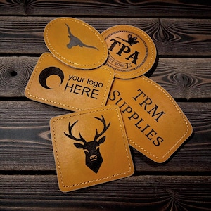 以下が含まれることがあります： さまざまな黒いデザインとテキストが施された、タン色の革製パッチのコレクション。パッチには、ロングホーンのシルエット、「TPA EST. 2025」のテキストが入った円形ロゴ、「your logo HERE」の三日月、鹿の頭、「TRM SUPPLIES」が含まれています。