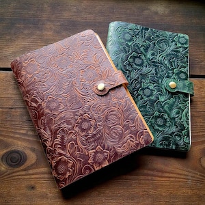 Könnte beinhalten: Zwei Lederjournale mit geprägten Blumenmustern. Eines ist braun, das andere dunkelgrün. Beide haben einen Riemenverschluss mit einem goldfarbenen Knopf. Die Journale werden auf einer Holzoberfläche präsentiert.