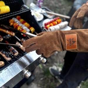 Może przedstawiać: Brązowa skórzana rękawica do grillowania z logo "Daddy's BBQ Gloves". Rękawica jest używana do trzymania szczypiec nad grillem z jedzeniem w trakcie gotowania.
