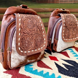 May include: Two brown leather backpacks with intricate floral tooled designs. The backpacks have a white cowhide patch on the front and are sitting on a colorful rug.
