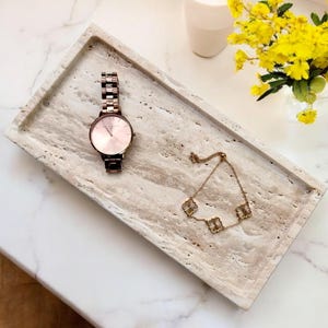May include: A rectangular, beige travertine tray holds a rose gold watch and a gold bracelet with clover-shaped charms. The watch has a pink face and a metal band. Yellow flowers in a small vase are in the background.