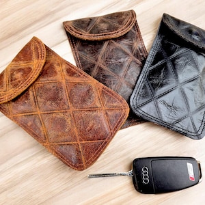 Könnte beinhalten: Drei Leder-Handyhüllen mit gestepptem Design. Die Hüllen sind braun, schwarz und bräunlich. Ein schwarzer Autoschlüssel mit einem silbernen Schlüsselring befindet sich im Vordergrund.
