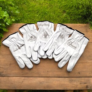 May include: Five pairs of white leather gardening gloves with personalized names and logos. The gloves have black trim and are laid out on a wooden surface.