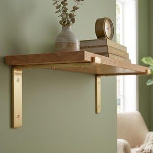 Puede incluir: Una repisa de madera sostenida por dos soportes metálicos en forma de L de color oro cepillado. La repisa sostiene un pequeño jarrón de cerámica con tallos de eucalipto secos y una pila de dos libros con un reloj analógico redondo de marco dorado.