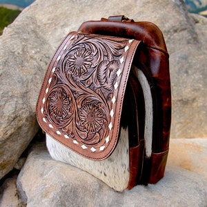 May include: A brown leather backpack with a white and brown cowhide flap. The flap has a floral design embossed into the leather. The backpack is sitting on a rock.