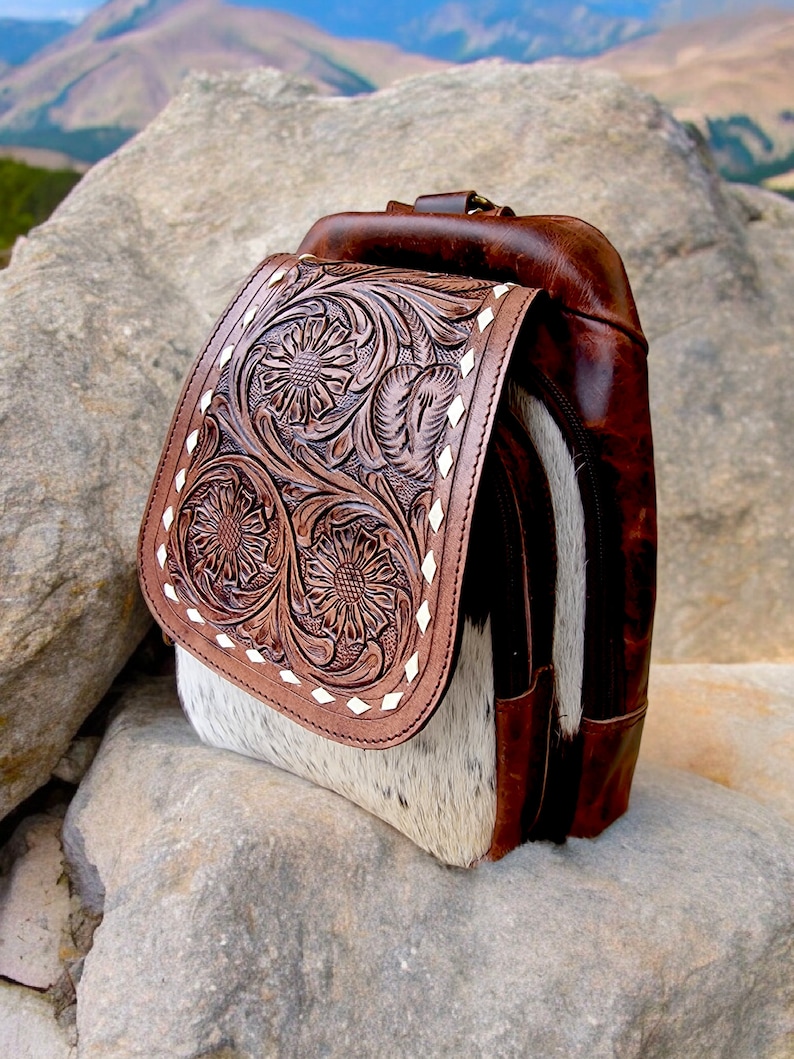 May include: A brown leather backpack with a white and brown cowhide flap. The flap has a floral design embossed into the leather. The backpack is sitting on a rock.