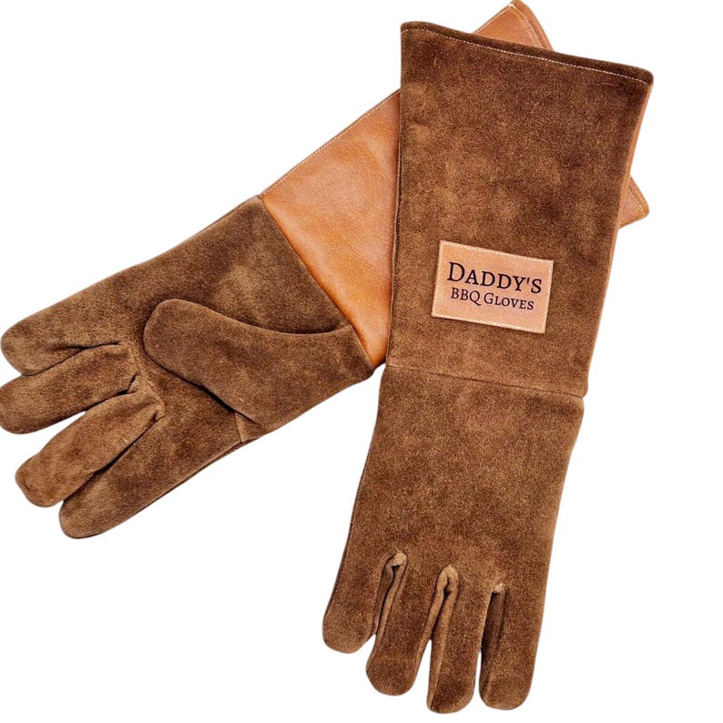 K&ouml;nnte beinhalten: Ein Paar braune Leder-Grillhandschuhe mit einem hellbraunen Lederflicken auf der R&uuml;ckseite des linken Handschuhs. Der Flicken tr&auml;gt den Text "Daddy's BBQ Gloves" in dunkelvioletter Schrift.