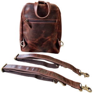May include: A brown leather backpack with two adjustable straps. The backpack has a zippered pocket on the front and two metal rings on the sides.