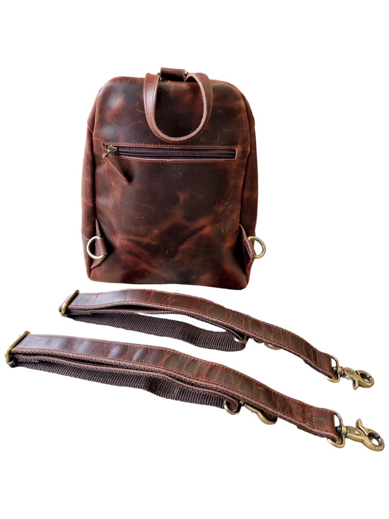 May include: A brown leather backpack with two adjustable straps. The backpack has a zippered pocket on the front and two metal rings on the sides.
