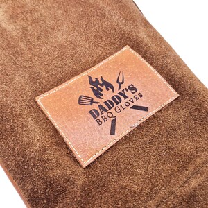 K&ouml;nnte beinhalten: Braune Leder-Grillhandschuhe mit einem braunen Leder-Patch, auf dem "Daddy's BBQ Gloves" mit einer Flamme und Grillwerkzeug-Grafik steht.