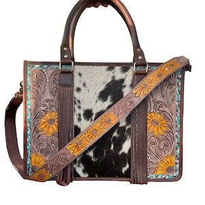 Puede incluir: Un bolso de mano de cuero marrón con un patrón de piel de vaca en blanco y negro. El bolso tiene una correa larga y un asa superior. El bolso está decorado con bordados de girasoles y detalles turquesa.