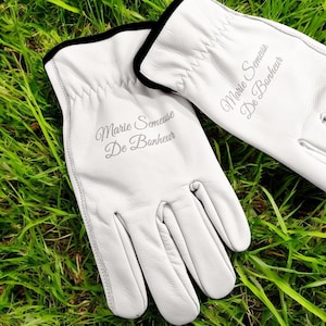 May include: A pair of white leather gloves with black trim. The gloves are embroidered with the text "Marie Semeuse De Bonheur".
