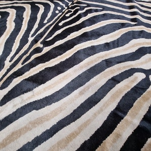 Pode incluir: Uma imagem de perto de um tapete com listras de zebra. O tapete apresenta listras pretas, bege e branco sujo alternadas, criando um padr&atilde;o arrojado de estampa animal. A textura do tapete parece macia e felpuda, adequada para uma sala de estar ou quarto.