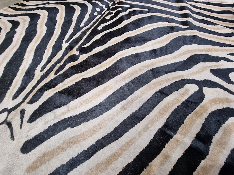 Pode incluir: Uma imagem de perto de um tapete com listras de zebra. O tapete apresenta listras pretas, bege e branco sujo alternadas, criando um padr&atilde;o arrojado de estampa animal. A textura do tapete parece macia e felpuda, adequada para uma sala de estar ou quarto.
